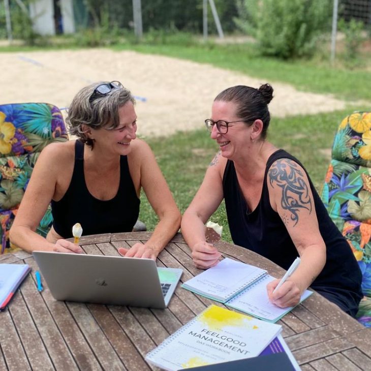 Zwei Frauen lachen und arbeiten an einem Tisch im Freien, umgeben von Natur.